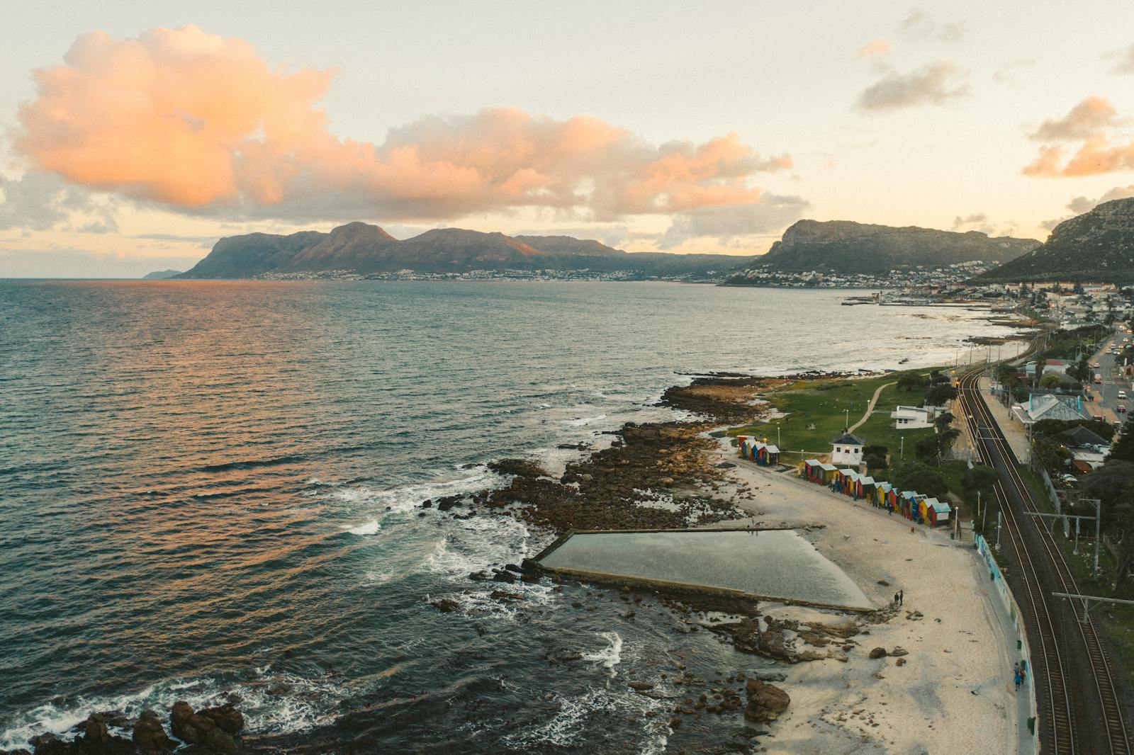 Stunning aerial view of a coastal city with colorful beach huts, mountains, and ocean at sunset.